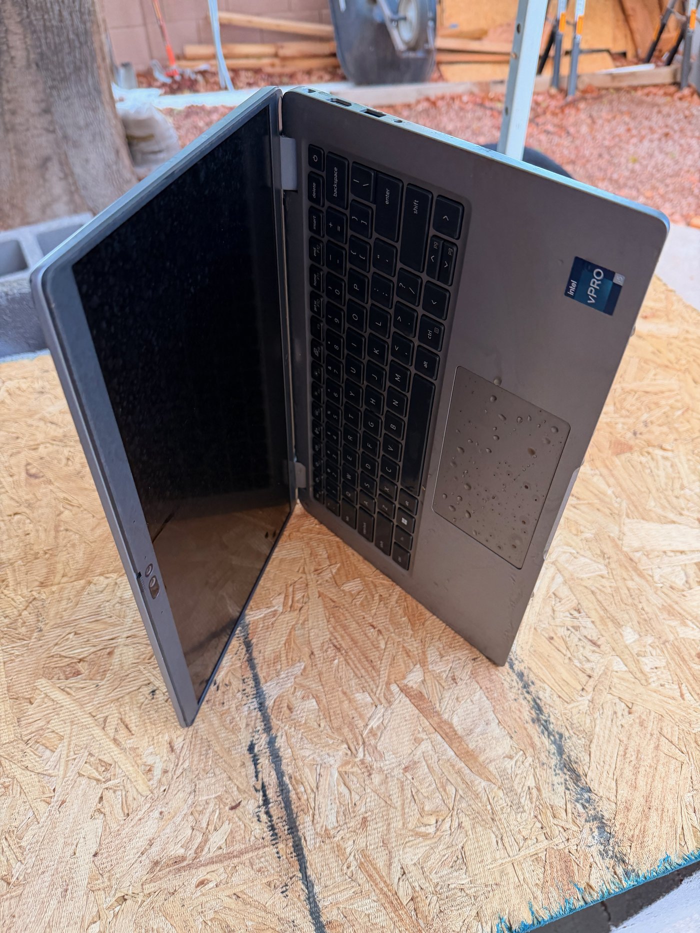 Work laptop propped open outdoors on plywood, its keyboard beaded with rainwater